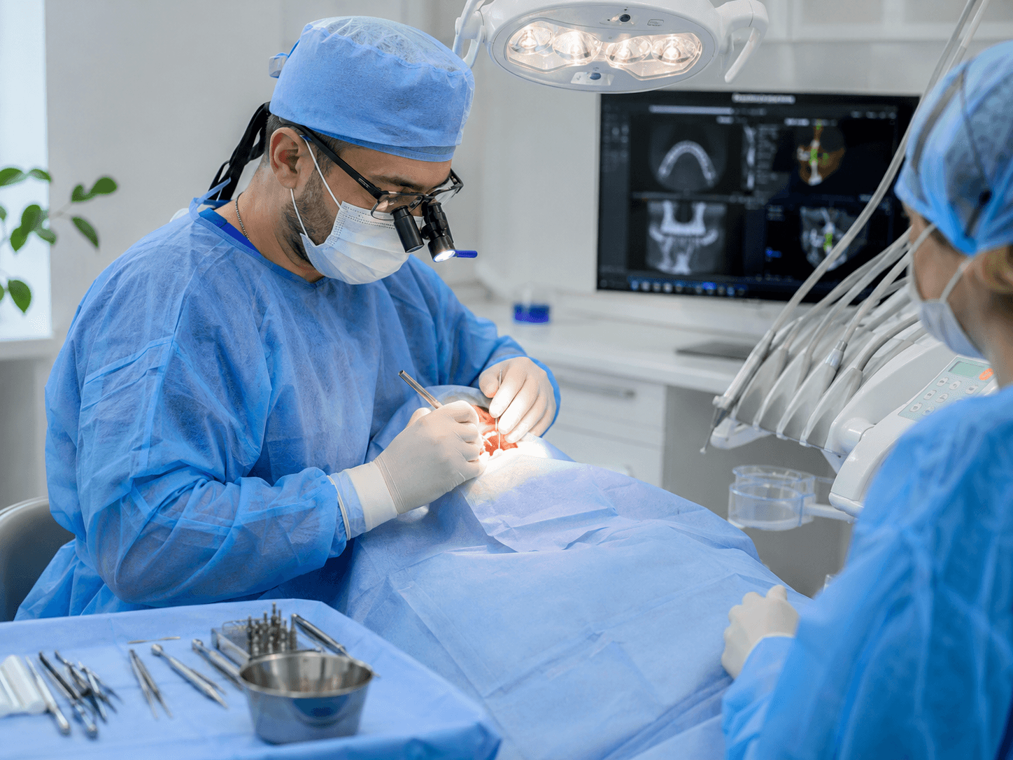수술복과 마스크를 착용한 치과의사가 confocal 조명 아래에서 임플란트 시술 도구를 들고 환자를 치료하는 모습, 뒤편 모니터에는 CT 영상이 표시됨